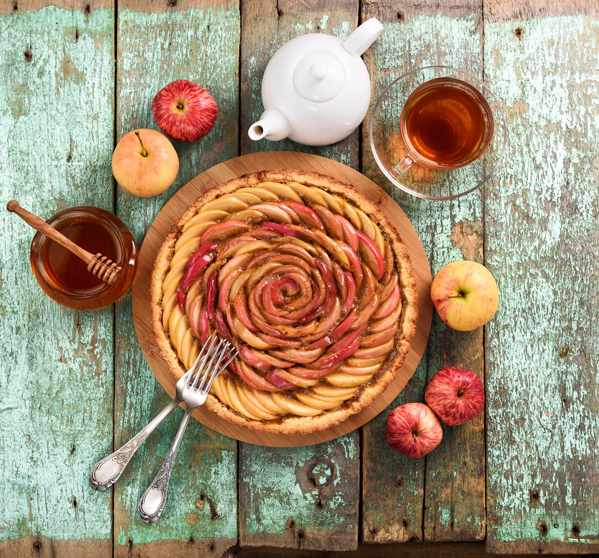 Tartă trandafir cu mere și miere pe un platou de lemn, alături de mere, un borcan cu miere și o ceașcă de ceai