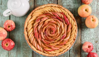 Tartă trandafir cu mere și miere pe un platou de lemn, alături de un ceainic alb și mai multe mere