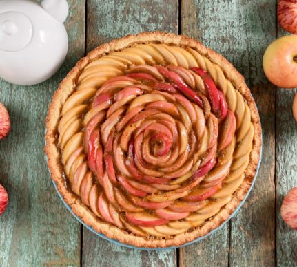 Tartă trandafir cu mere și miere pe un platou de lemn, alături de un ceainic alb și mai multe mere
