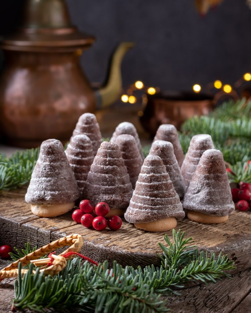 11 brăduți de Crăciun din biscuiți cu sos de ciocolată, pe o masă rustică de lemn, alături de un ceainic vitange într-un decor de sărbătoare