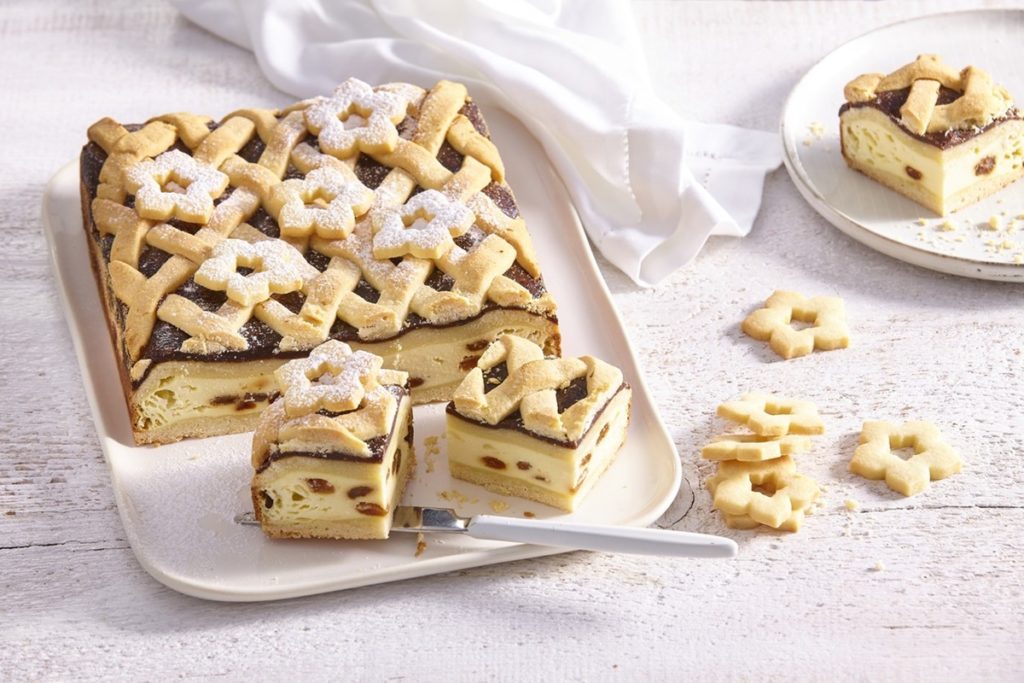 Prăjitură fragedă cu brânză și gem de prune porționată pe platou și pe farfurie, alături de o sită cu zahăr pudră și biscuiți