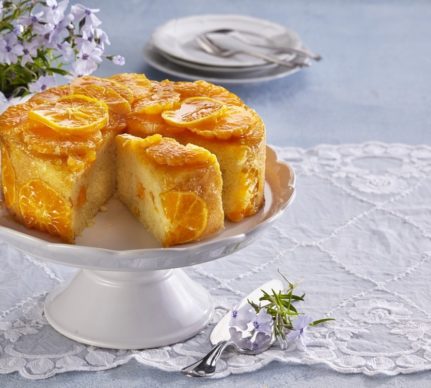 Tort cu mandarine porționat pe un platou cu picior, alături de o vază cu flori și farfurii cu tacâmuri de servit