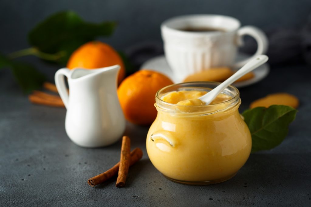 Orange curd în borcan cu linguriță, alături de o ceașcă cu cafea, o zaharniță, portocale și batoane de vanilie
