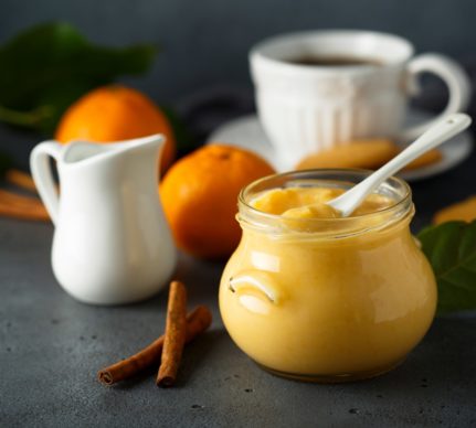 Orange curd în borcan cu linguriță, alături de o ceașcă cu cafea, o zaharniță, portocale și batoane de vanilie