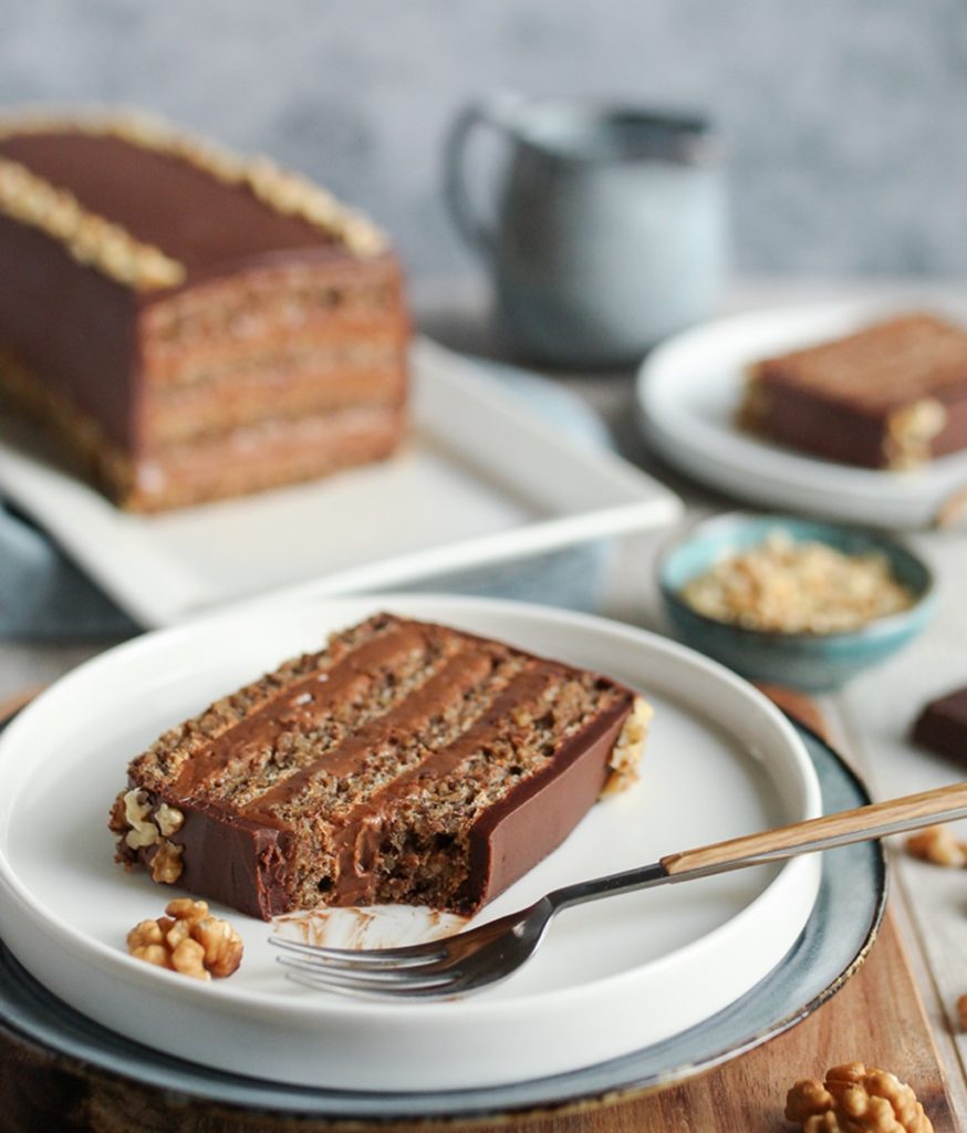 Două porții de tort pe farfurii albe, una secționată cu furculița, alături de platoul cu tort, un bol cu alune și ciocolată mărunțită