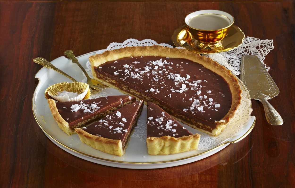 Tartă cu ciocolată și cremă caramel cu migdale porționată pe platou, alături de două lingurițe, o spatulă și o ceașcă cu farfuriuță