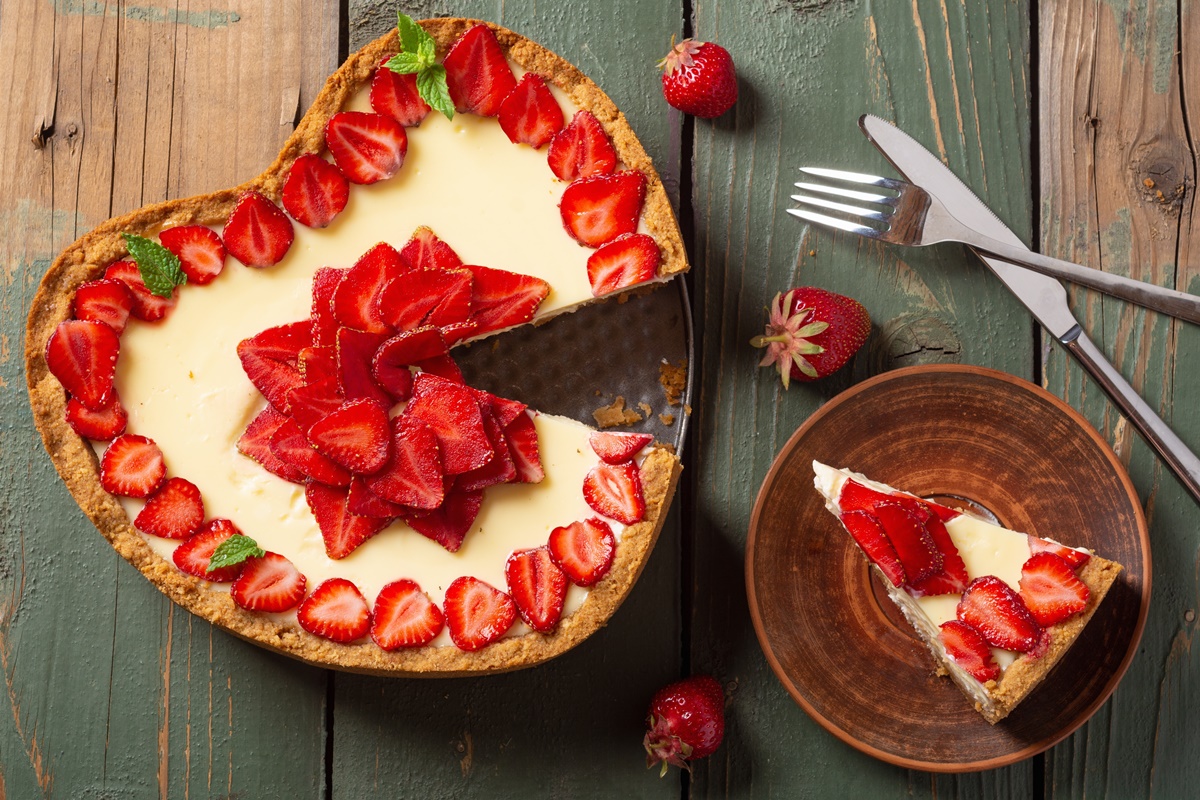 Cheesecake inimioară cu căpșuni secționat pe platou și o porție pe farfurie, alături de un cuțit și o furculiță