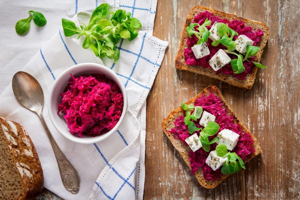 Cremă de sfeclă roșie în bol și pe felii de pâine, cu bucăți de brânză și frunze de salată