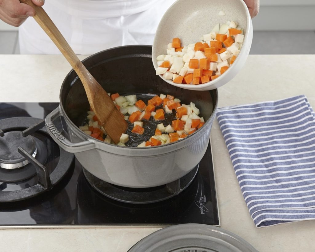 Adăugare morcovi cu țelină din bol alb în cratiță cu spatulă de lemn