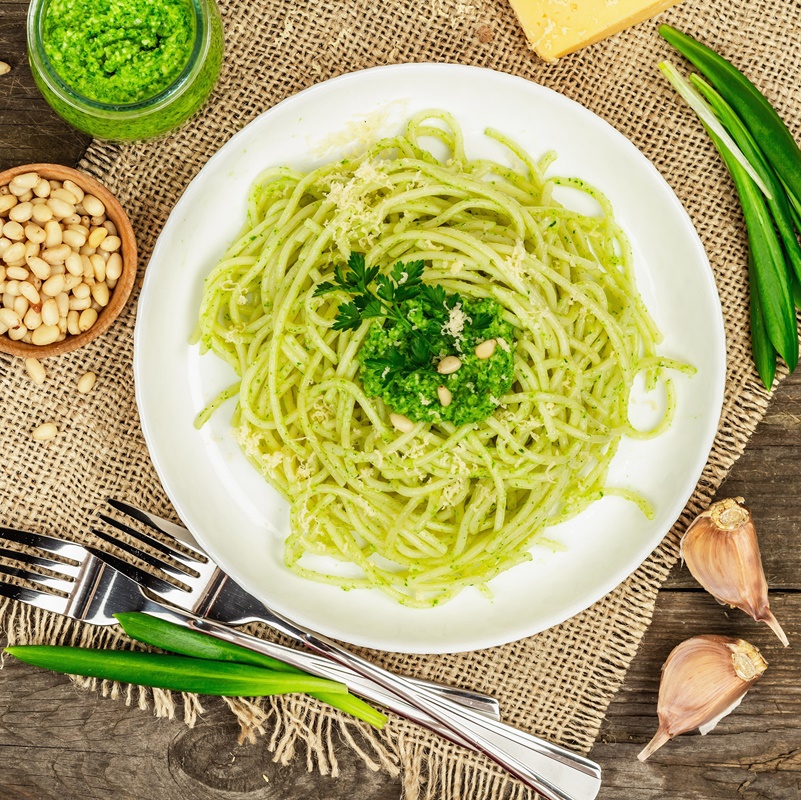 Spaghete cu pesto de leurdă și parmezan în farfurie albă cu două furculițe, frunze de leurdă, un bol cu semințe de pin, un borcan cu sos pesto și o bucată de parmezan