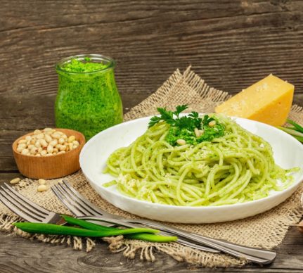 Spaghete cu pesto de leurdă și parmezan în farfurie albă cu două furculițe, frunze de leurdă, un bol cu semințe de pin, un borcan cu sos pesto și o bucată de parmezan