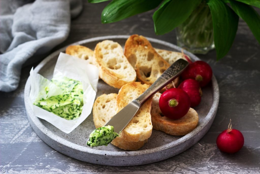 Unt aromat cu leurdă, pătrunjel și ceapă verde pe cuțit și pe hârtie, alături de felii de pâine și ridichi