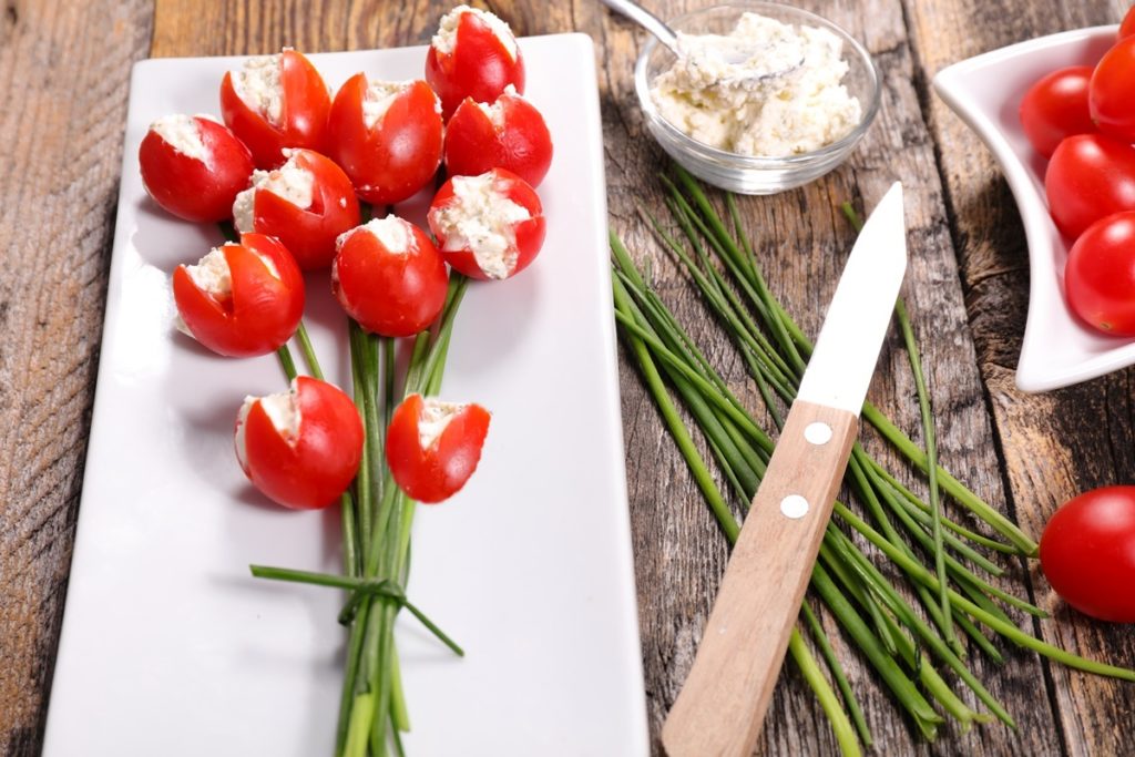 Aperitiv lalele din roșii cu brânză și ceapă verde pe un platou alb, alături de un bol cu brânză și linguriță, un bol cu roșii, cozi de ceapă și un cuțit