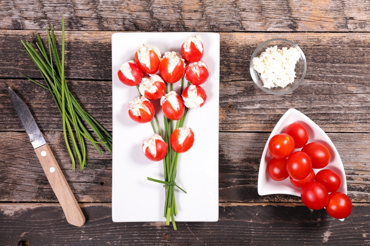 Aperitiv lalele din roșii cu brânză și ceapă verde pe un platou alb, alături de un bol cu brânză și linguriță, un bol cu roșii, cozi de ceapă și un cuțit