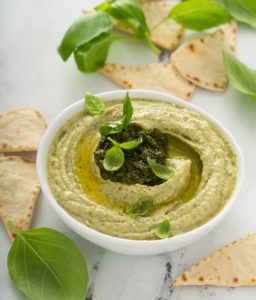 Hummus cu pesto de busuioc în bol alb, alături de lipii și frunze proaspete de busuioc