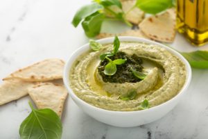 Hummus cu pesto de busuioc în bol alb, alături de lipii și frunze proaspete de busuioc