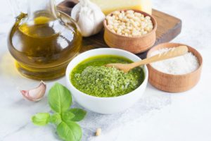 Sos pesto de busuioc în bol alb, alături de o sticlă cu ulei, un bol cu semințe de pin, sare și frunze de busuioc