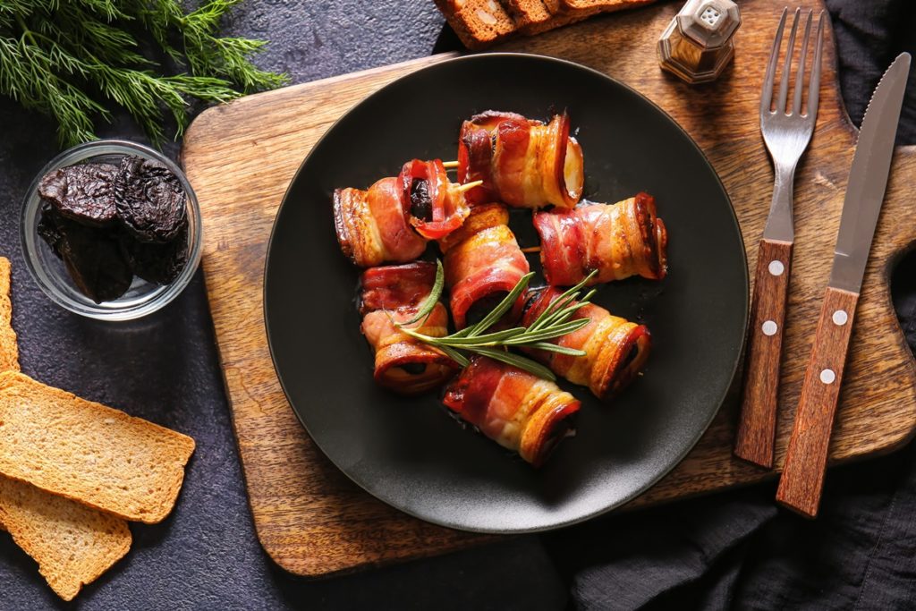 Rulouri de bacon cu prune uscate, decorate cu rozmarin, pe o farfurie neagră