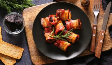 Rulouri de bacon cu prune uscate, decorate cu rozmarin, pe o farfurie neagră