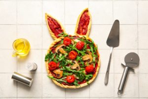 Tartă sub formă de iepuraș, cu ciuperci, roșii cherry și rucola, alături de o spatulă, un cuțit de pizza, un recipient cu ulei și solnițe