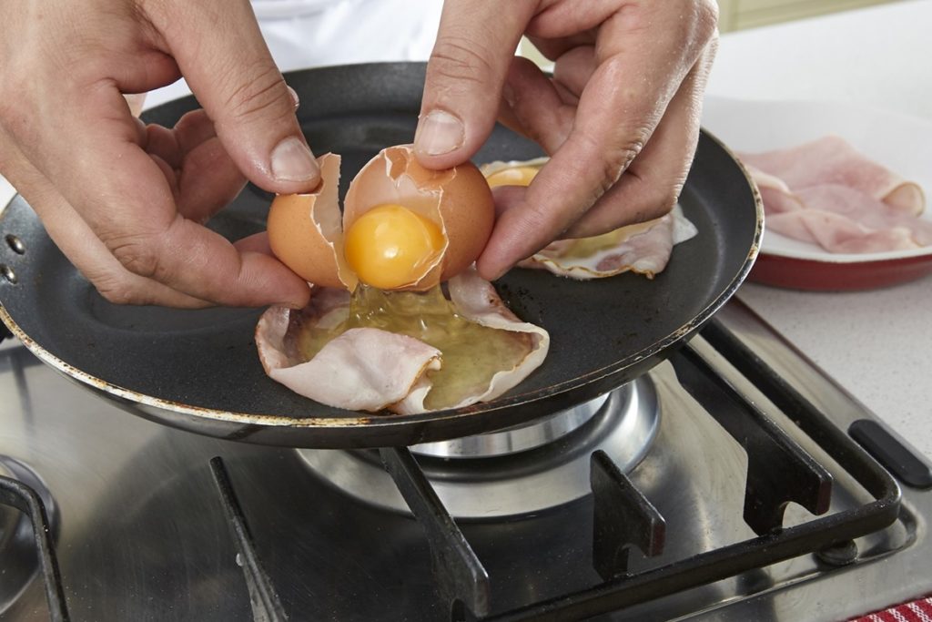 Persoană adăugând oul în coșulețul de bacon din tigaie