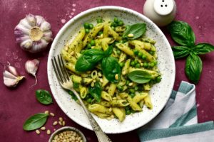 Penne cu sos pesto și mazăre într-o farfurie albă cu furculiță, alături de o solniță, o căpățână de usturoi, un bol cu semințe de pin și frunze de busuioc