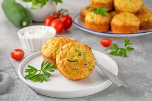 Doi muffins cu dovlecei și verdețuri pe o farfurie, alături de un platou cu mai mulți muffins, un bol cu sos și roșii proaspete