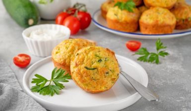 Doi muffins cu dovlecei și verdețuri pe o farfurie, alături de un platou cu mai mulți muffins, un bol cu sos și roșii proaspete