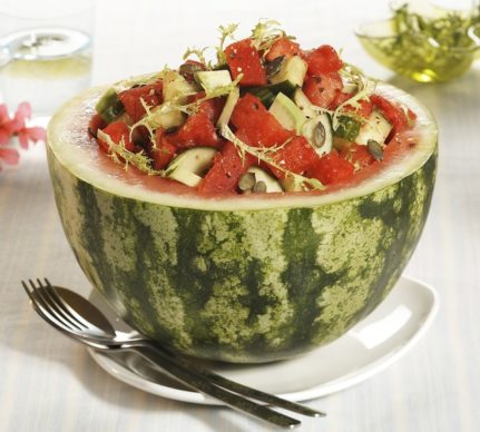 Salată de pepene roșu cu avocado și castraveți, decorată cu frunze de andive și semințe, în cupă de pepene roșu, alături de lingură și furculiță
