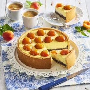 Cheesecake cu mac și caise proaspete porționat pe platou și o porție pe farfurie, alături de două cești de cafea și caise proaspete