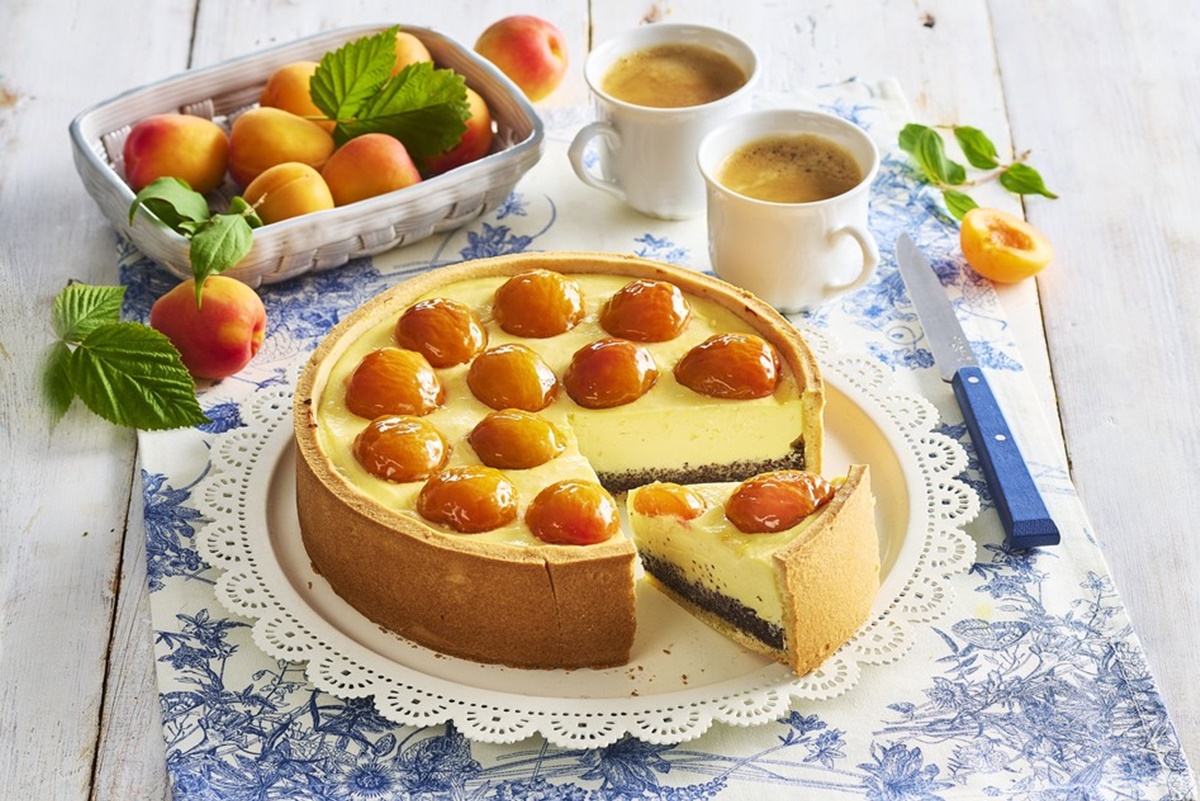 Cheesecake cu mac și caise proaspete porționat pe platou, alături de un bol cu caise și două cești de cafea