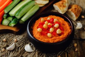 Porție de hummus cu ardei copți în bol negru, alături de legume proaspete și lipii