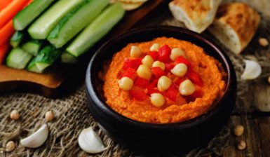 Porție de hummus cu ardei copți în bol negru, alături de legume proaspete și lipii