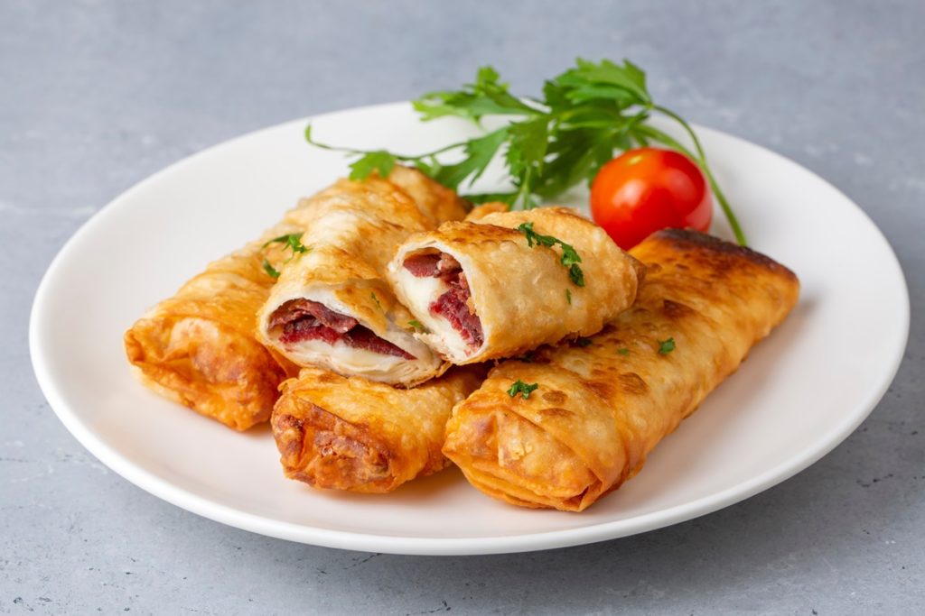 Patru porții de Borek cu pastramă și brânză cheddar, una secționată pe platou, alături de o roșie și pătrunjel verde