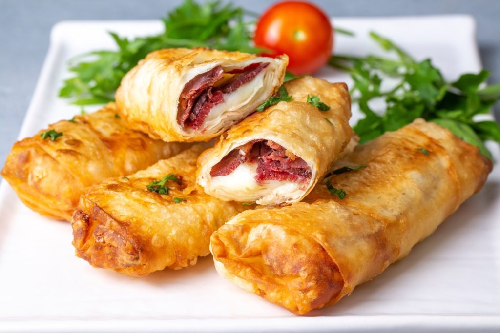 Patru porții de Borek cu pastramă și brânză cheddar, una secționată pe platou, alături de o roșie și pătrunjel verde