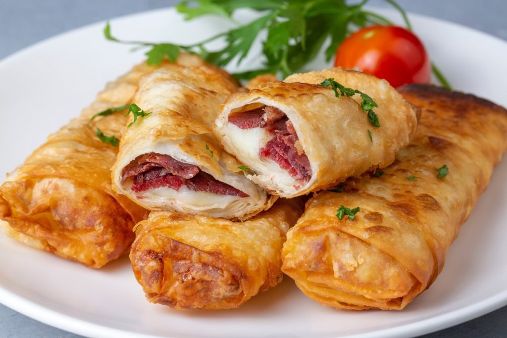 Patru porții de Borek cu pastramă și brânză cheddar, una secționată pe platou, alături de o roșie și pătrunjel verde