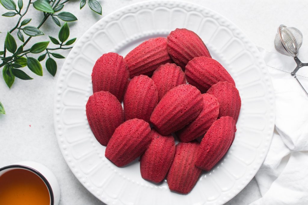 Madeleine Red Velvet pe un platou alb, alături de o sită cu zahăr și o ceașcă cu ceai