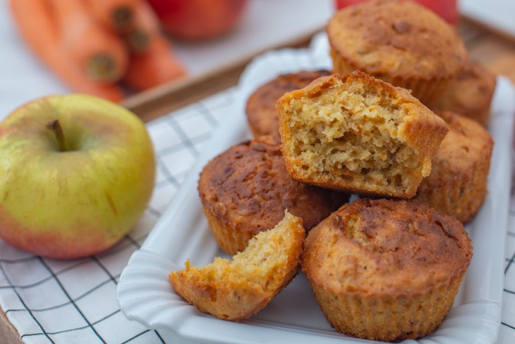 Muffins cu morcovi și mere pe un platou alb, alături de mere proaspete și morcovi