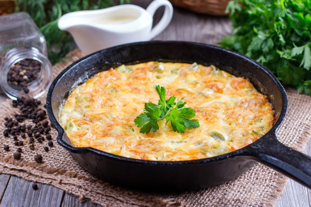 Budincă cu praz și brânză în tigaie, alături de un bol cu smântână, pătrunjel verde și un borcan cu piper
