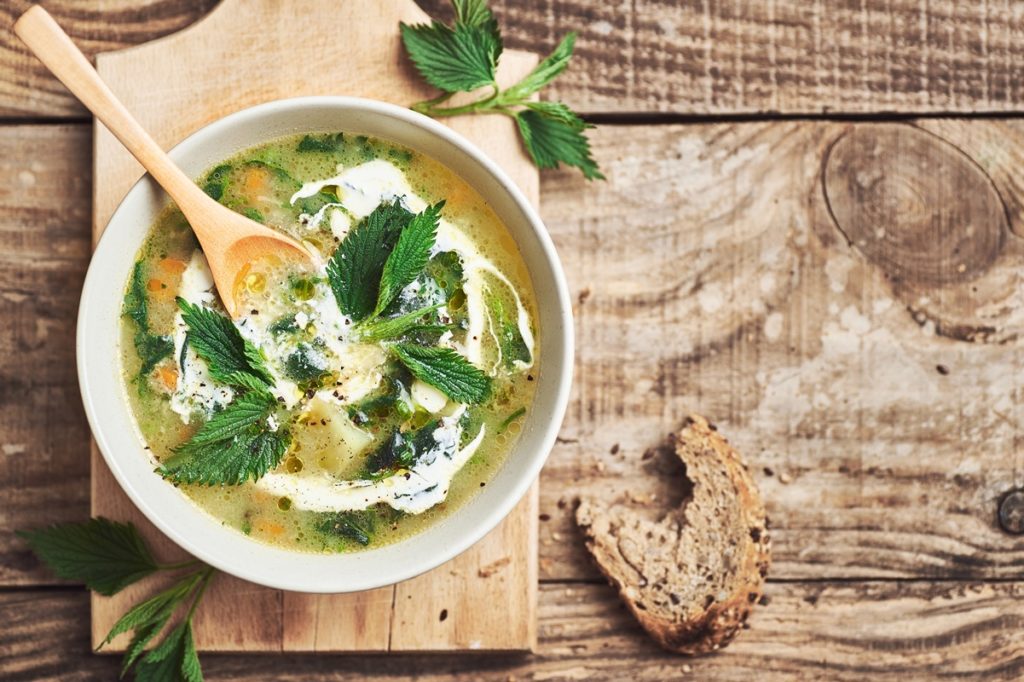 Ciorbă din urzici în bol alb cu lingură pe un tocător de lemn, alături de o felie de pâine ruptă