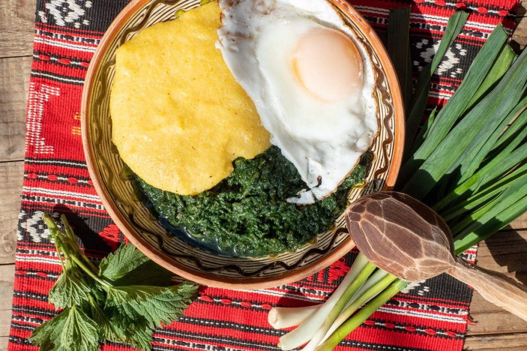 Mâncare de urzici cu ouă ochiuri și mămăligă în farfurie, alături de lingură de lemn, ceapă verde și frunze de urzici pe un ștergar rustic