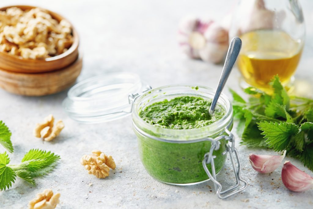 Pesto de urzici cu nuci în borcan, alături de un bol cu nuci, o sticlă cu ulei, urzici și usturoi