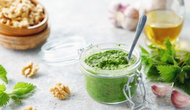 Pesto de urzici cu nuci în borcan, alături de un bol cu nuci, o sticlă cu ulei, urzici și usturoi