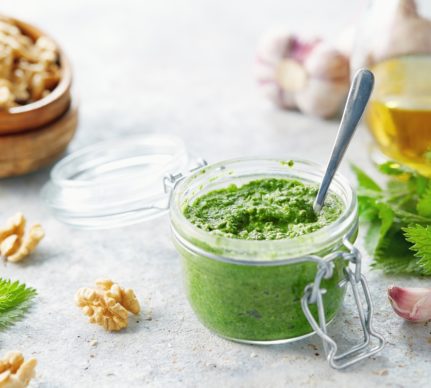 Pesto de urzici cu nuci în borcan, alături de un bol cu nuci, o sticlă cu ulei, urzici și usturoi