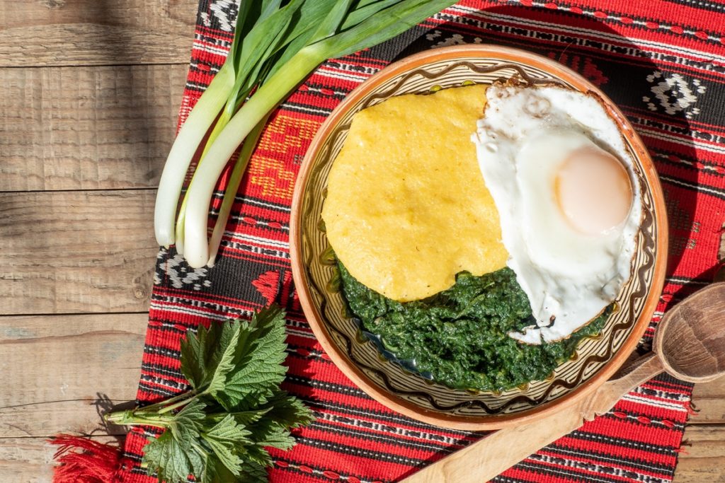 Mâncare de urzici cu ouă ochiuri și mămăligă în farfurie, alături de ceapă verde, lingură de lemn și frunze de urzici pe un ștergar rustic