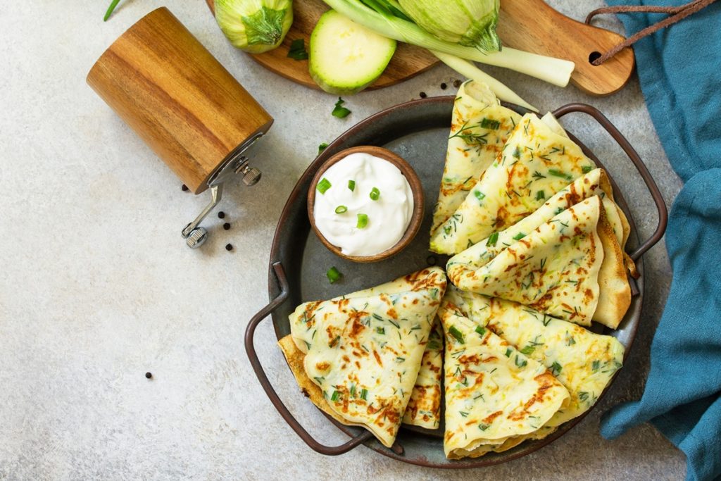 Clătite de dovlecei cu verdețuri de primăvară pe un platou, alături de un bol cu iaurt, o râșniță, dovlecei și ceapă verde