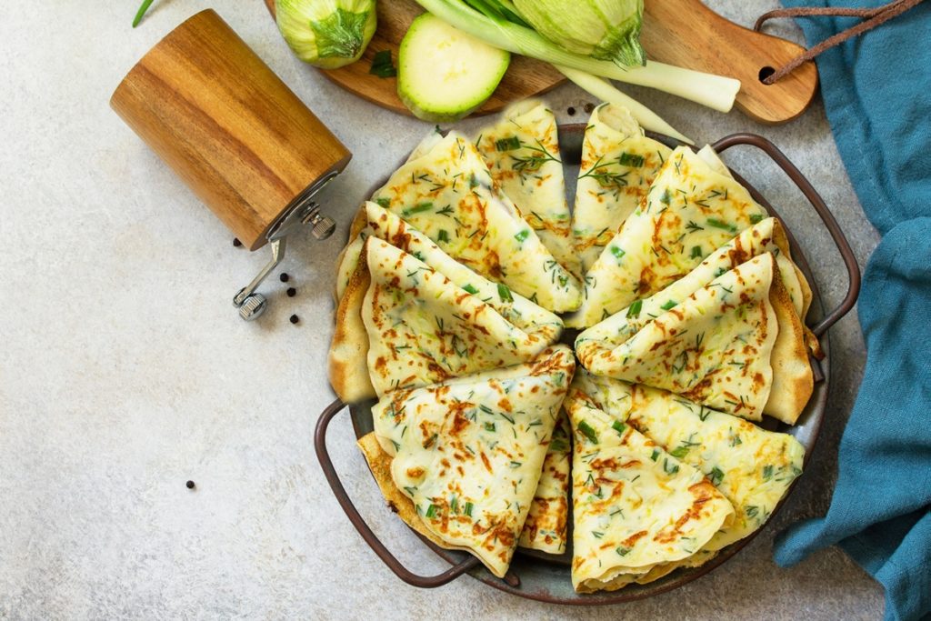 Clătite subțiri din dovlecei cu verdețuri de primăvară pe un platou, alături de o râșniță, dovlecei și ceapă verde