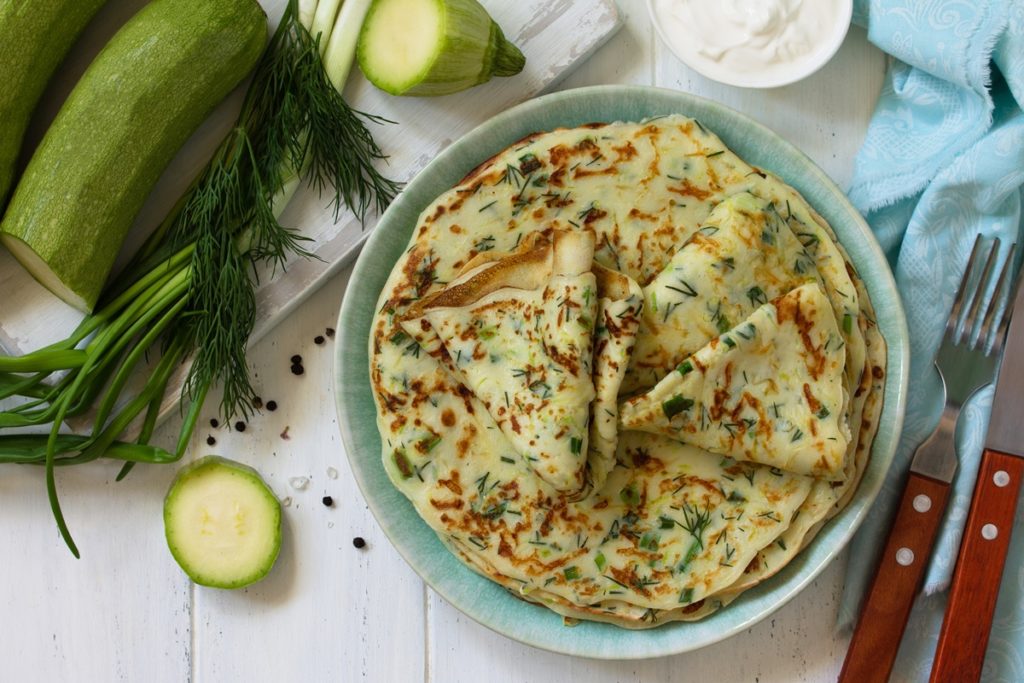 Clătite subțiri din dovlecei cu verdețuri de primăvară pe un platou, alături de furculiță și cuțit, un bol cu iaurt, dovlecei, mărar și ceapă verde