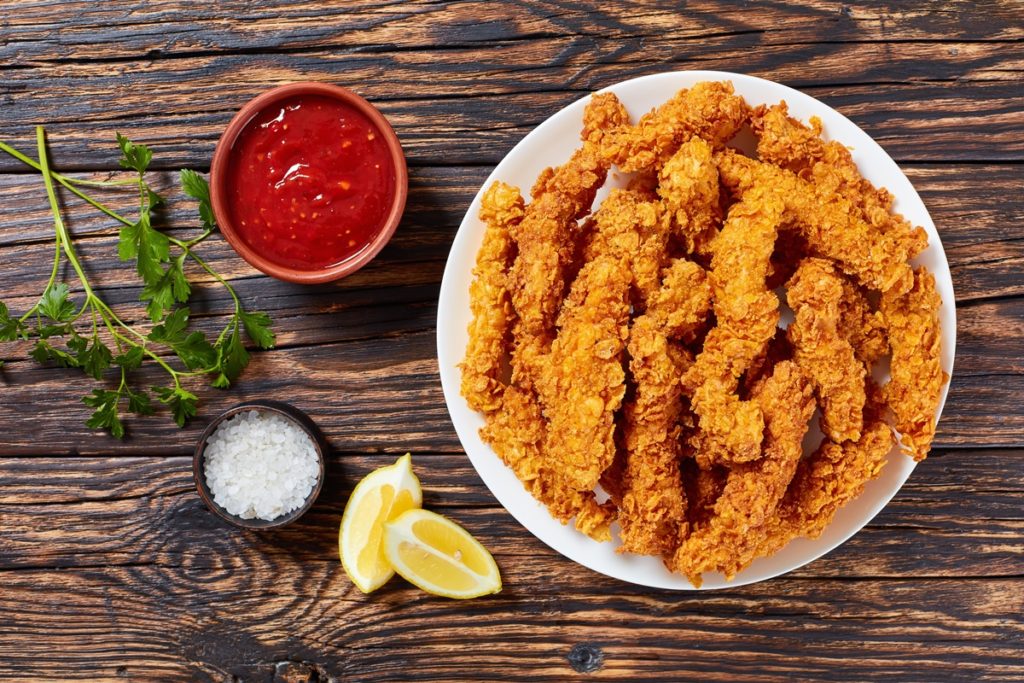 Degețele din piept de pui în crustă de porumb pe un platou, alături de un bol cu sos de roșii, unul cu sare și două felii de lămâie