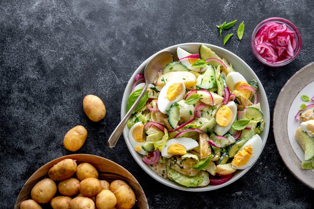Salată de cartofi cu ceapă roșie și ouă fierte în bol alb, alături de un bol cu ceapă roșie murată, un bol cu cartofi și unul cu verdeață